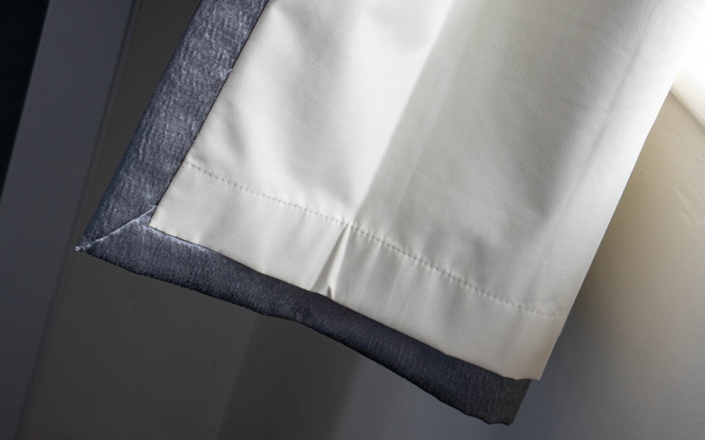 Close-up of the bottom edge of a Couture Living curtain in a bedroom, featuring a white inner fabric with grey outer lining, illuminated by natural light from a nearby window.