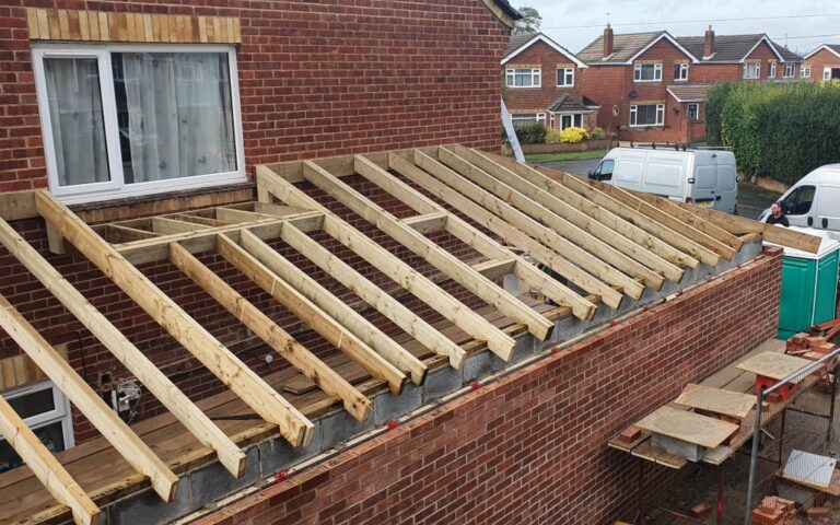 A house extension with exposed wooden roof beams and red brick walls is under construction, featuring a roof extension in progress, scaffolding, and vans parked nearby on a residential street.
