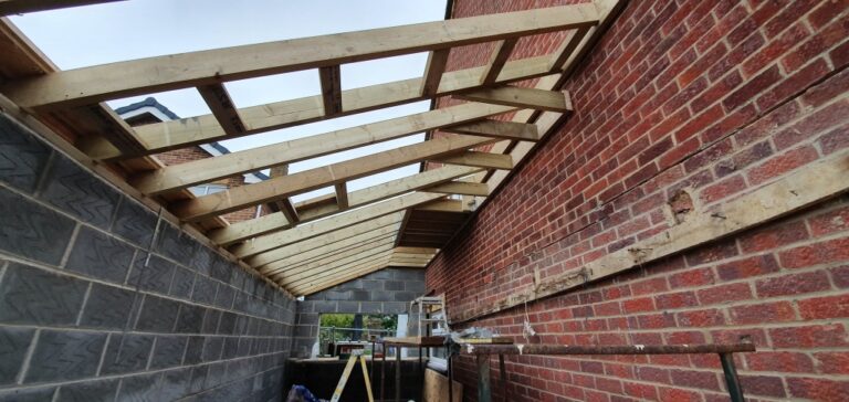 Wooden roof rafters are being installed on a house extension between two brick walls; construction materials and a ladder are visible underneath the incomplete roof structure.