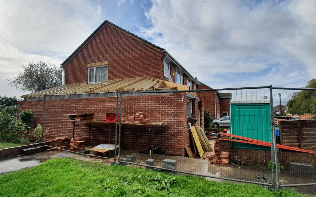 A brick house extension is underway, with wooden beams forming the framework for a new roof extension. Building materials and bricks are stacked by a metal fence. Nearby, a green shed stands under the cloudy sky.