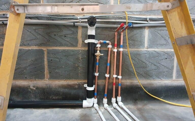 Copper and plastic pipes, some with blue and red tape, are attached to a wall with grey blocks—possibly part of a house extension. A shut-off valve is in the centre, framed by yellow ladders, with a yellow cable visible on the right.