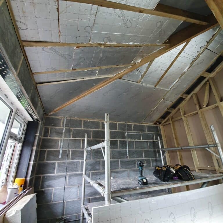 A room under construction for a house extension with exposed brickwork, wooden studs, visible cables and pipes, partially insulated ceiling panels, scaffolding, a drill, and a toolbag. Natural light enters through a window on the left.