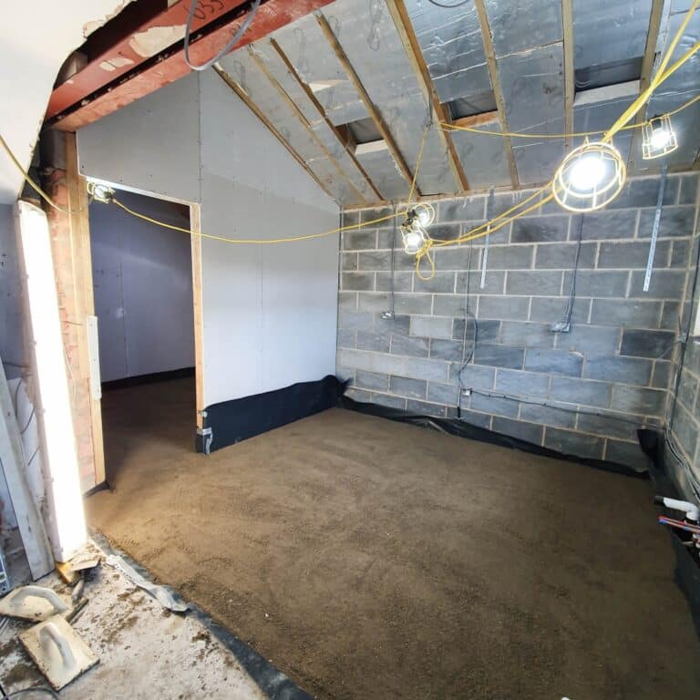 A room under construction shows clear building progress, with exposed brick walls, an unfinished ceiling, hanging work lights, and a floor covered in sand or gravel. Tools and materials for the home refurbishment are visible around the edges.