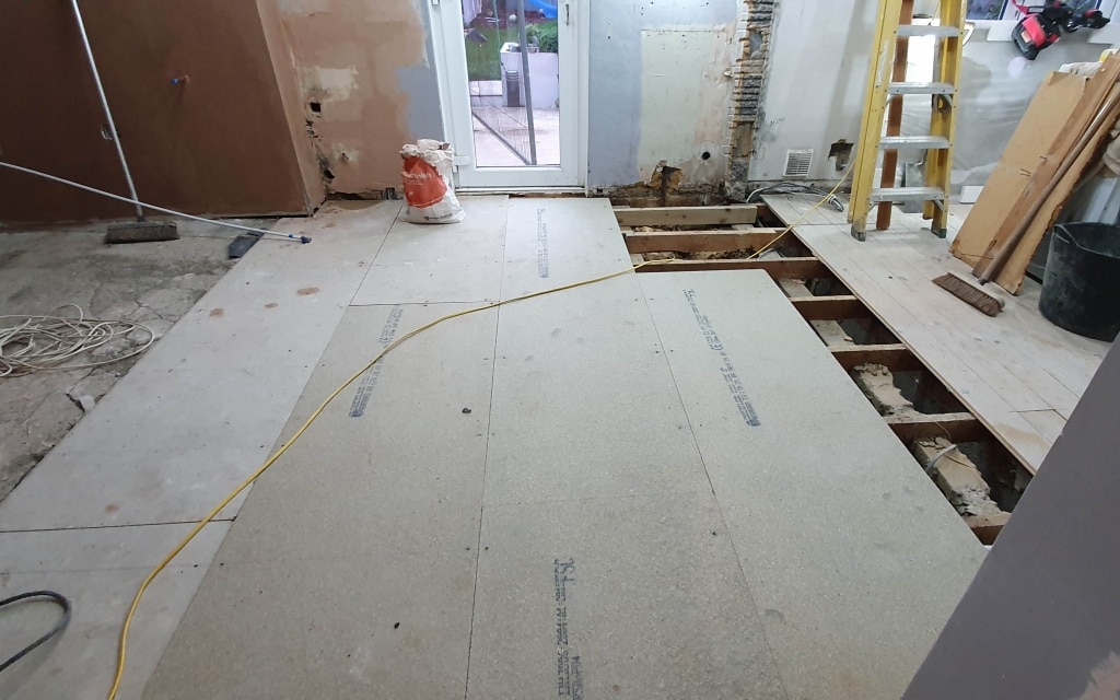 A partially renovated room shows progress in the house extension: some new floorboards fitted, exposed floor joists, building materials, a yellow ladder, and tools scattered about. The door at the back leads outside.