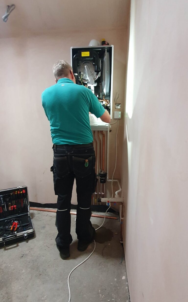 A person in a teal shirt and work trousers is servicing a wall-mounted boiler in a room, with tools nearby—essential maintenance to keep things running smoothly during an extension week of the house extension project.