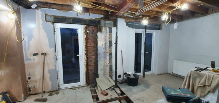 A room under home renovation with exposed brickwork, unfinished walls, and construction tools scattered on the floor. Two glass doors lead outside, while bright work lights hang from the ceiling—marking progress in the week 11 house extension.