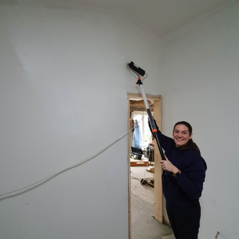 A smiling person uses a long-handled paint roller to paint a white wall in a room under renovation during week 12 of the house extension, with another room visible through a doorway in the background.