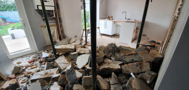 A partially demolished interior wall shows walls coming down, with rubble and debris scattered across the floor; structural supports hold up the ceiling, and a kitchen area is visible in the background—suggesting a house extension in progress.