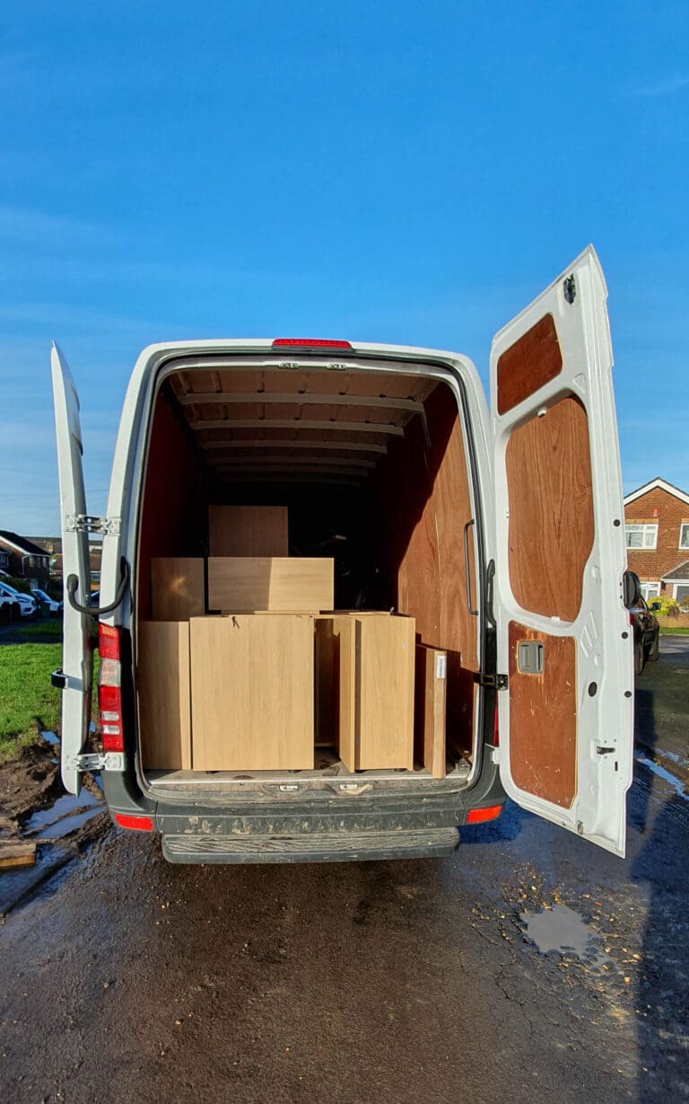 A white van with rear doors open reveals several light wooden cupboards, possibly for a house extension, stacked inside. The van is parked on a residential street under a clear blue sky.