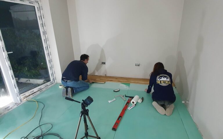 Two people are installing wooden flooring in a house extension. Tools and materials, including a camera on a tripod, a spirit level, and boards, are scattered around them. A large window is visible on the left side of the image.