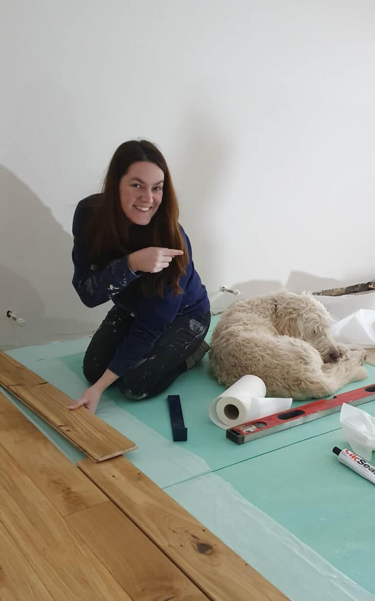 A woman kneels on the floor, smiling and pointing at a fluffy dog lying on its side. Surrounded by flooring tools and boards, they appear to be working on a house extension or home renovation project.