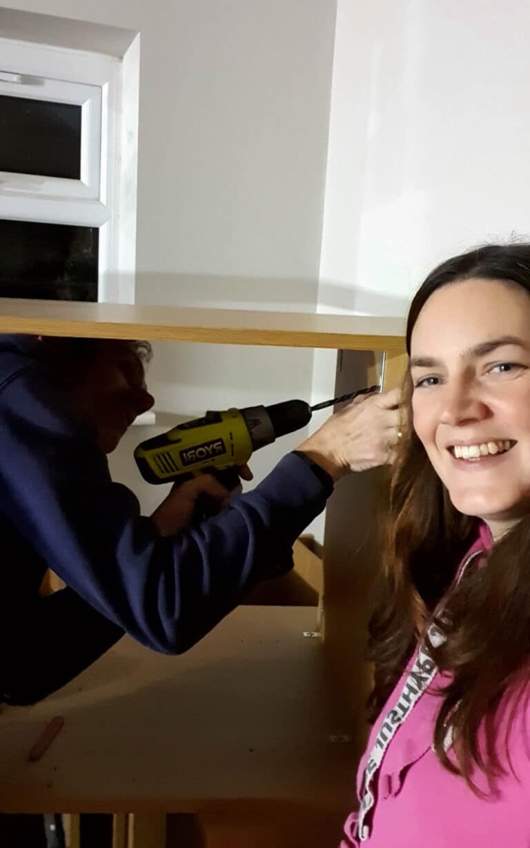 Two people assemble furniture indoors. One uses a yellow power drill while the other, in a pink top, smiles at the camera. A white wall and small window suggest they might be working on a house extension project together.