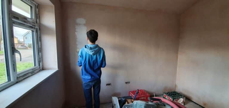 A person in a blue hoodie stands facing a bare wall with patched plaster in a house extension undergoing renovation, with tools and materials scattered on the floor near a large window.