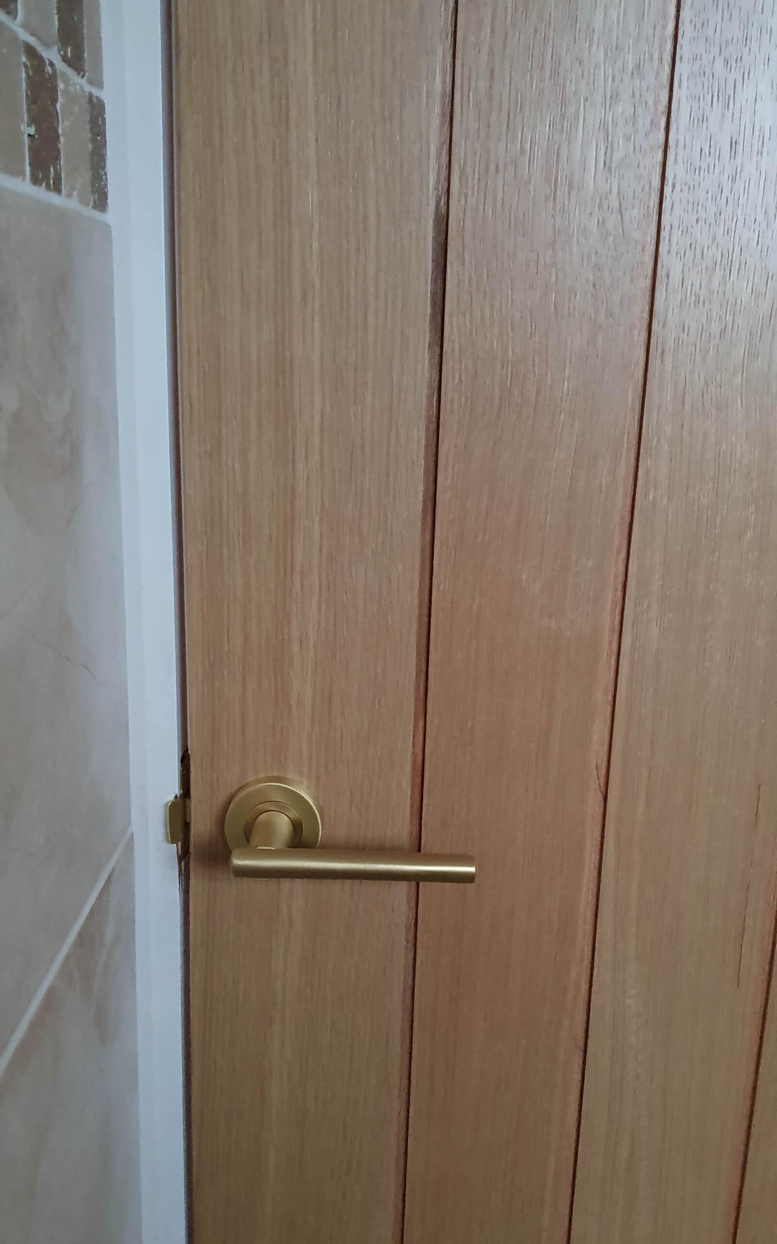 A wooden door with vertical panels and a gold-coloured handle, ideal for a room refurbishment. Beige tiled wall is visible on the left side of the image.