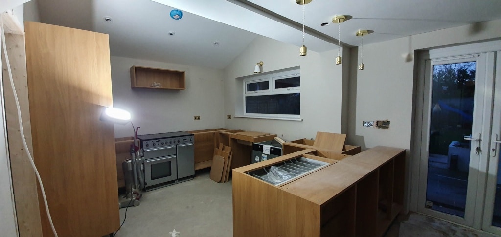 A kitchen under construction in a new house extension, with wooden cupboards being installed, an oven in place, loose wiring, a work light, and unpainted walls. There’s a window, door to the outside, and no worktops fitted yet—room not done.