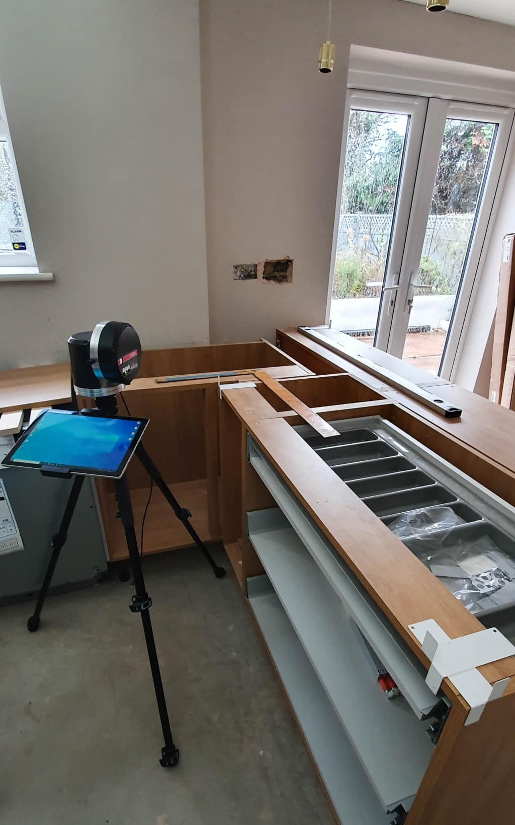 A partially assembled kitchen in a house extension features wooden cupboards, an open drawer, and a sink being fitted. A laser measuring device on a tripod and a tablet are set up nearby. Large windows show a garden outside as the room nears completion.