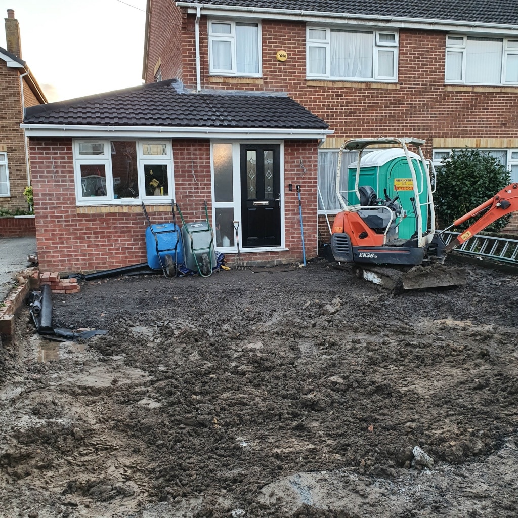 A muddy front garden is being prepared for landscaping or building work, possibly as part of a house extension, with a small orange digger, two wheelbarrows, and loose bricks in front of a brick house with white windows and doors.