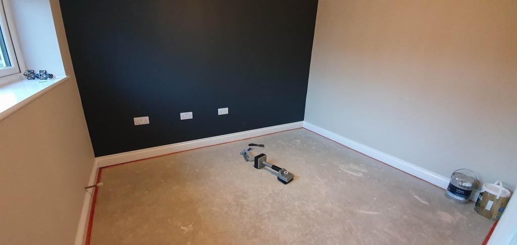 An empty room with a black feature wall, beige walls, and a window on the left is ready for home renovation. Painting supplies, paint tins, tools, and tape line the bare floor near the wall in preparation for updates.