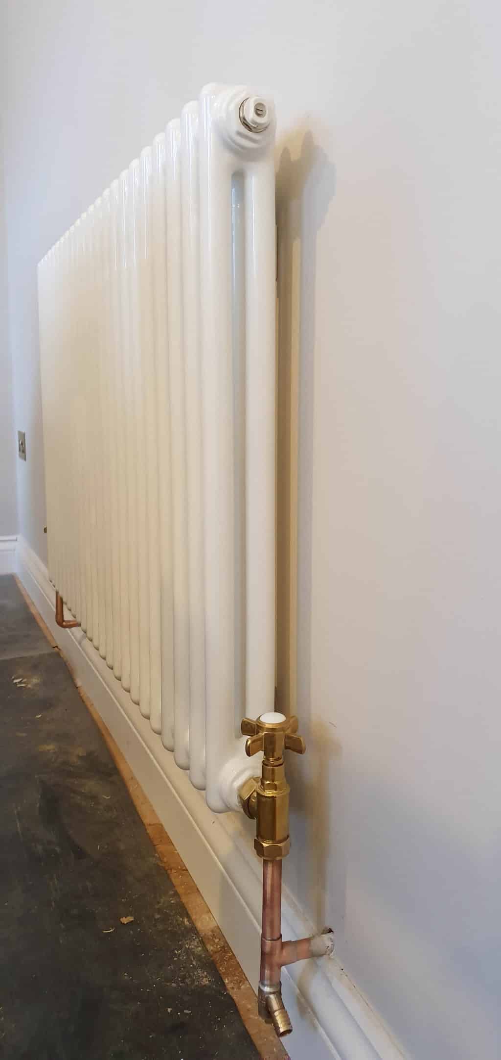 A white vertical column radiator attached to a light-coloured wall, with copper pipes and a valve at the bottom—an ideal choice for your next house extension, set beside a bare floor and skirting board.