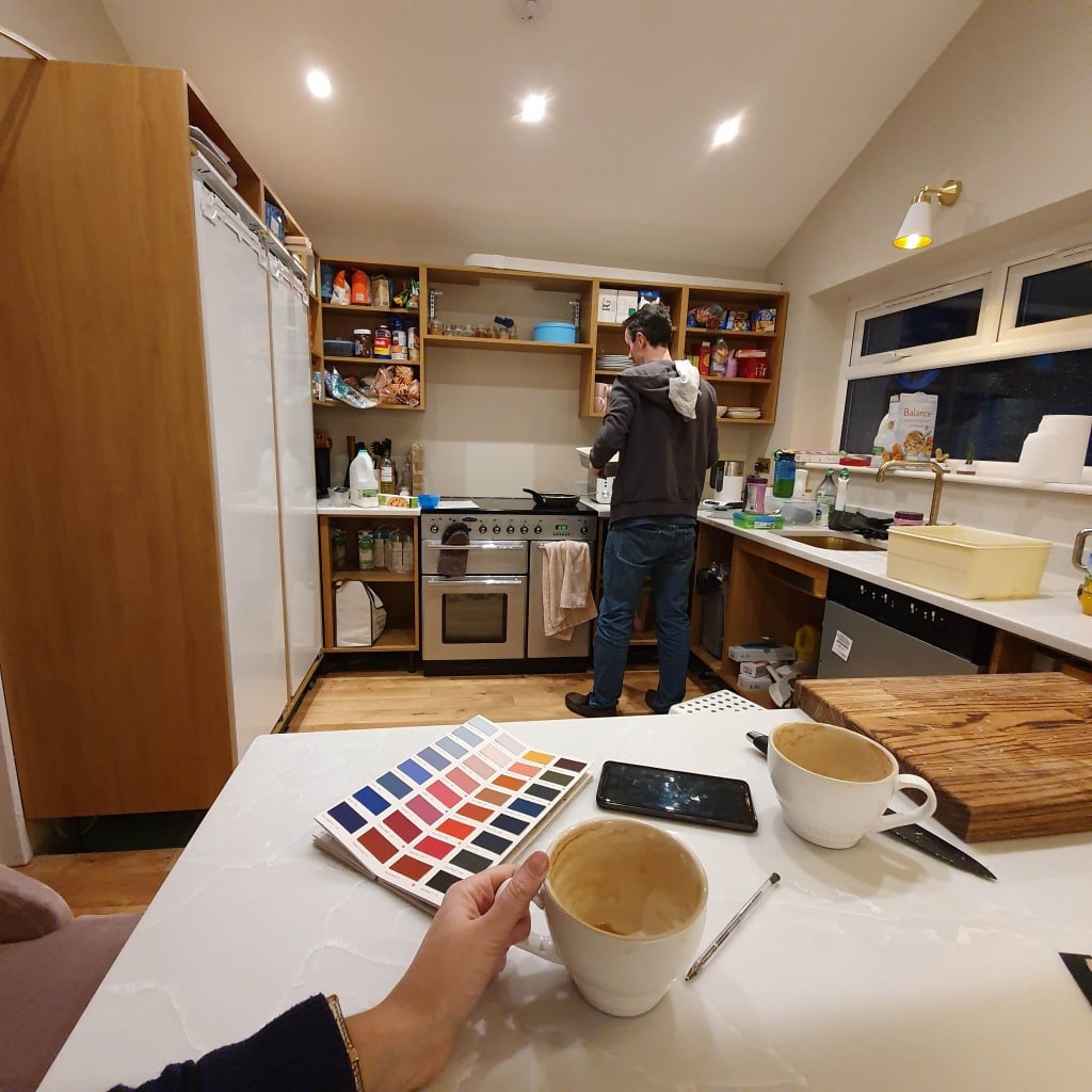 A person stands cooking in a cosy kitchen, possibly after a big refurbishment, while another hand holds a coffee cup and a colour swatch book at a white counter. Two mugs, a mobile, a pen, and chopping boards rest on the worktop.