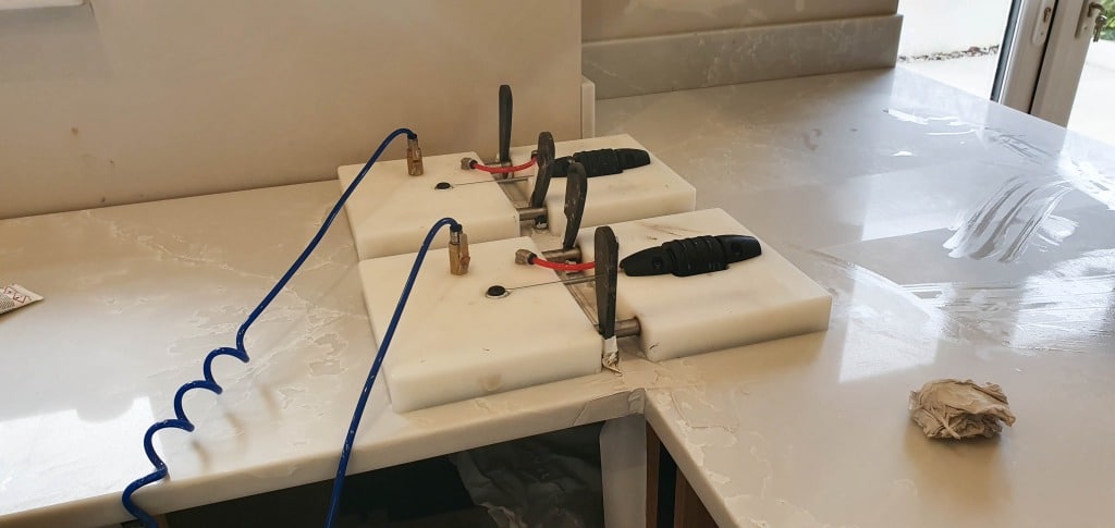 Two white suction clamps with black handles and a blue hose are securing and aligning two pieces of light-coloured worktop at a corner seam during a house extension installation. A kitchen roll sits on the right side of the worktop.