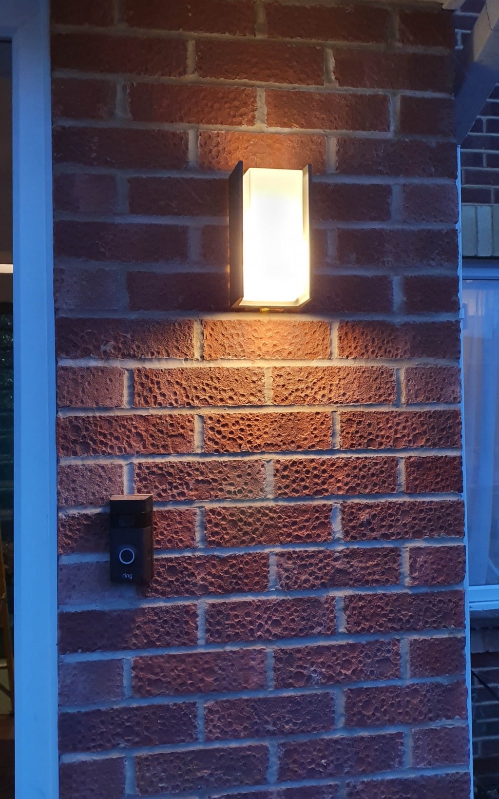 A modern wall light fitting emits a warm glow on a brick exterior wall above a black Ring video doorbell, highlighting the entrance to a home’s new house extension.