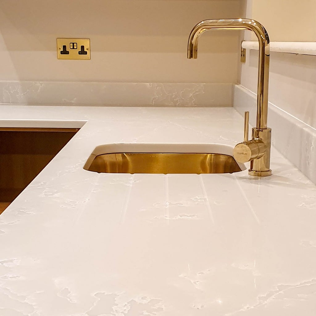 A modern kitchen worktop with a gold sink and matching gold tap, set in a white marble surface—a stunning big update perfect for any house extension. A power point is visible on the wall above the worktop.