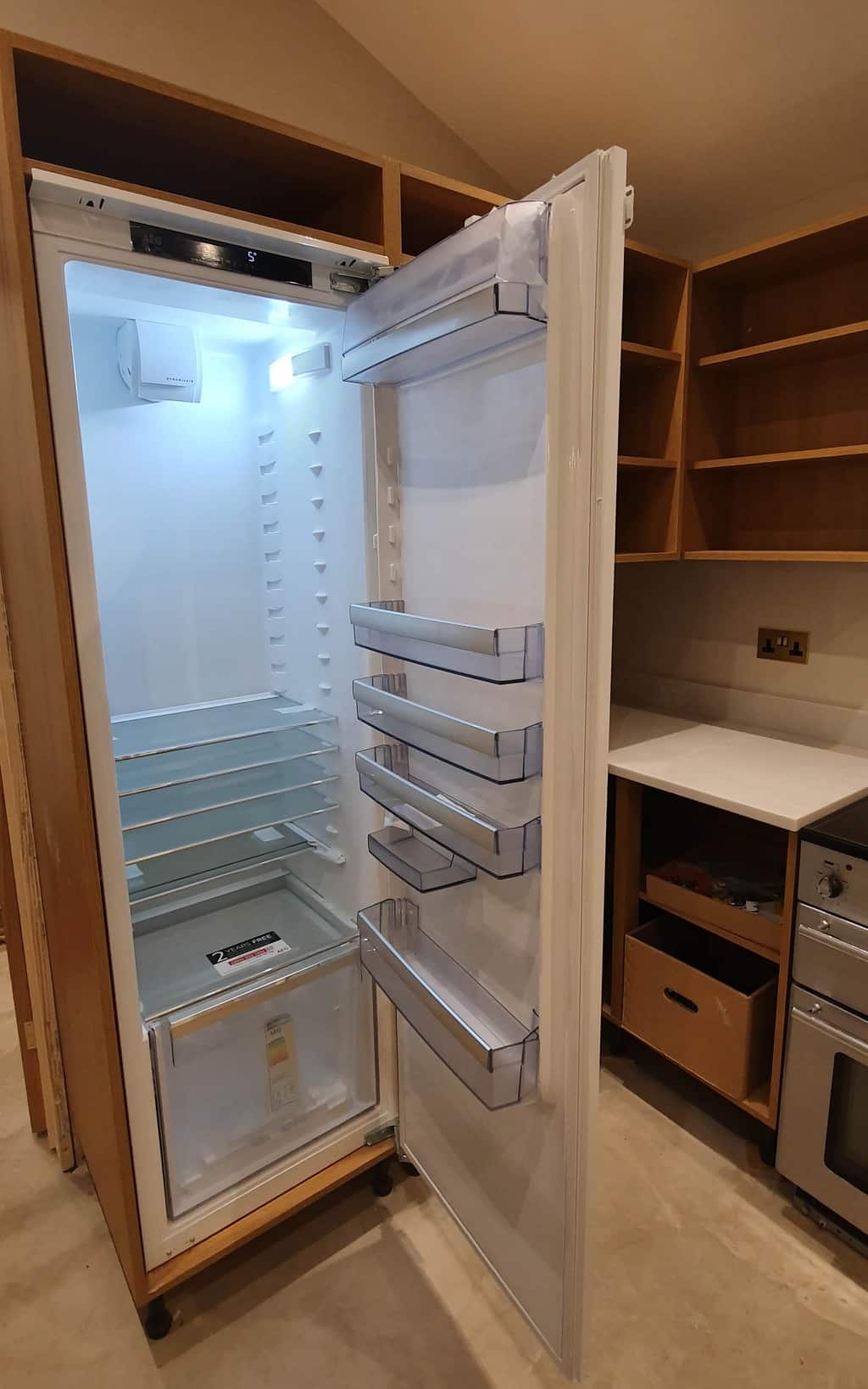 An empty fridge with the door open, showing clear shelves and door compartments, is built into wooden kitchen cabinetry. The surrounding kitchen, part of a major update and house extension, appears to be under construction or refurbishment.