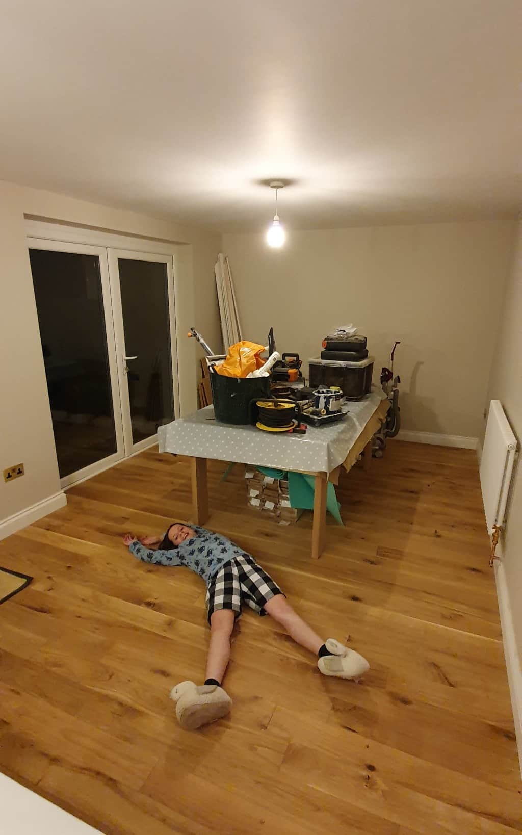 A person lies sprawled on a wooden floor in a mostly empty room. A table with household items sits in the background—perhaps part of a recent house extension, with bare walls and a large window brightening the space.