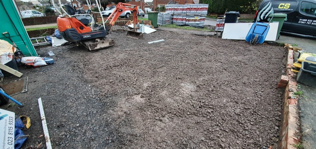 A building site with a small orange digger on a levelled earth area, surrounded by building materials and tools—looks like a major update for a house extension, with a van parked nearby on the residential road.