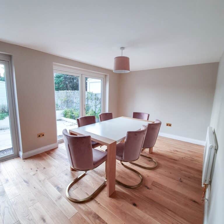 A bright dining room with a white rectangular table, six pink velvet chairs with gold legs, wooden flooring, beige walls, large windows, and a pink ceiling light—the perfect space after a big update or house extension.
