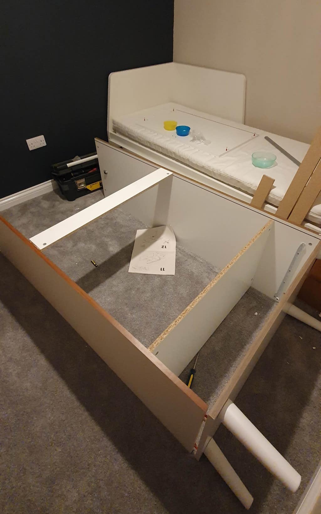 A partially assembled white bed frame lies on a grey carpet next to a matching bed with a mattress—tools, screws, an instruction sheet, and small containers are scattered around the room in the midst of a house extension.