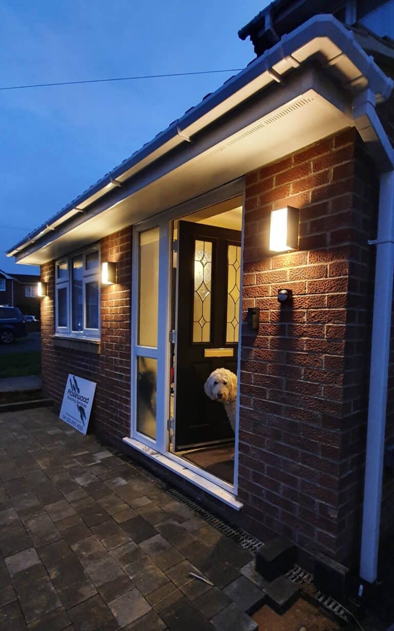 A warmly lit brick house with a house extension at dusk, its glass-panelled door open. A fluffy white dog looks out from inside whilst a sign is propped up against the house near the door.