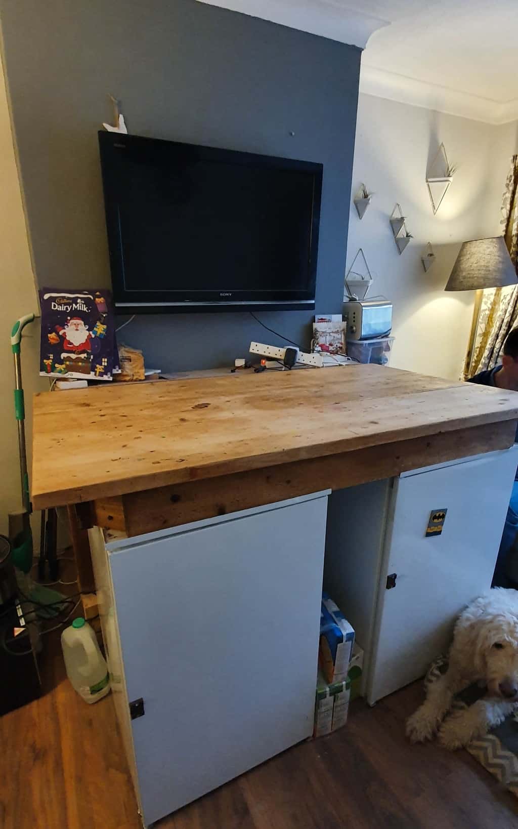 A large wooden workbench with a thick top stands against a grey wall beneath a mounted TV. White cupboards are underneath, with storage inside—part of a big update following the recent house extension. A dog lies on the floor next to the bench.