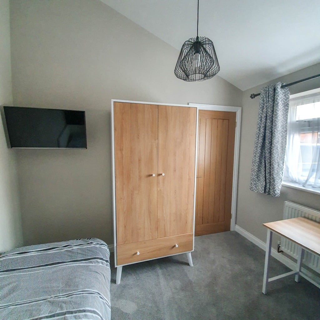 Bright, modern boys' bedroom with a striped bed, wooden wardrobe, wall-mounted TV, wooden door, and desk. Patterned curtains on the window and grey carpet create a stylish navy bedroom feel against the neutral walls.