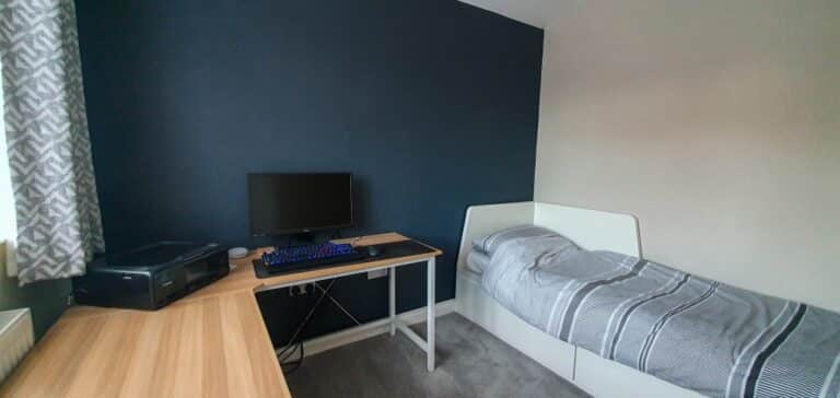 A neatly organised Boys’ Navy Bedroom with a single bed, grey bedding, a wooden desk holding a computer monitor, keyboard, mouse, and a printer by a window with patterned curtains. The wall behind the desk is dark blue.