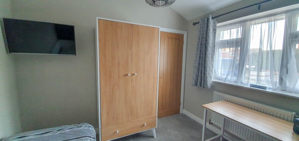 A tidy teenage boy’s bedroom with a wooden wardrobe, a wall-mounted TV, a bed partially visible, a desk by the window, and sheer curtains letting natural light brighten this navy bedroom.