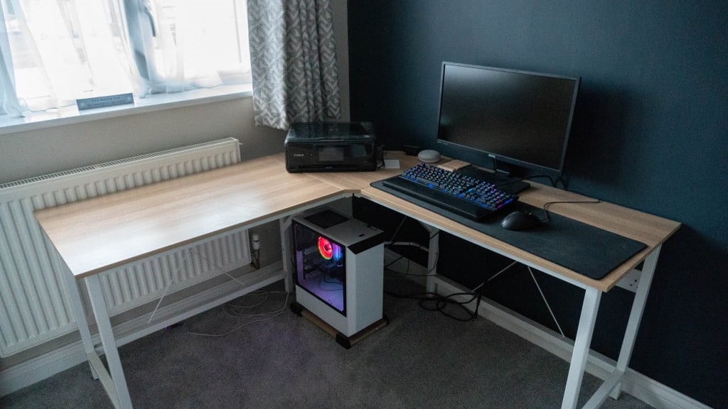 A tidy L-shaped desk with a monitor, keyboard, mouse, and printer on top. The setup sits in a carpeted boys' bedroom next to a window with white curtains, while a desktop computer with glowing lights rests underneath.