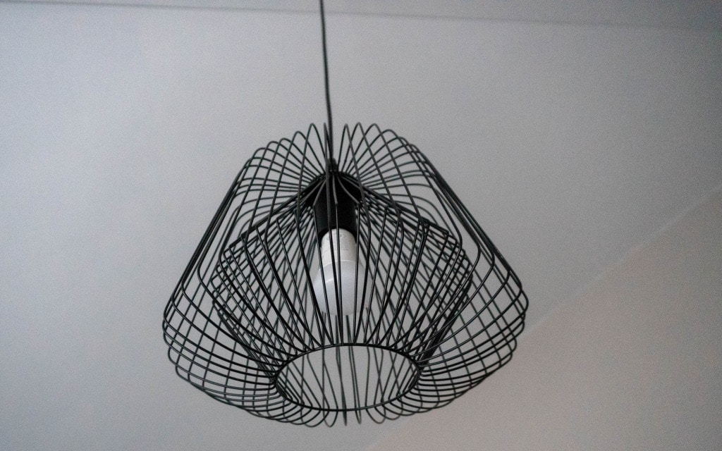 A black wire-frame lampshade with a geometric design hangs from the ceiling, surrounding a single exposed light bulb—an ideal accent for a teenage boys' bedroom or modern boys' navy bedroom with its bold, contemporary style. The background is a plain, light-coloured ceiling.