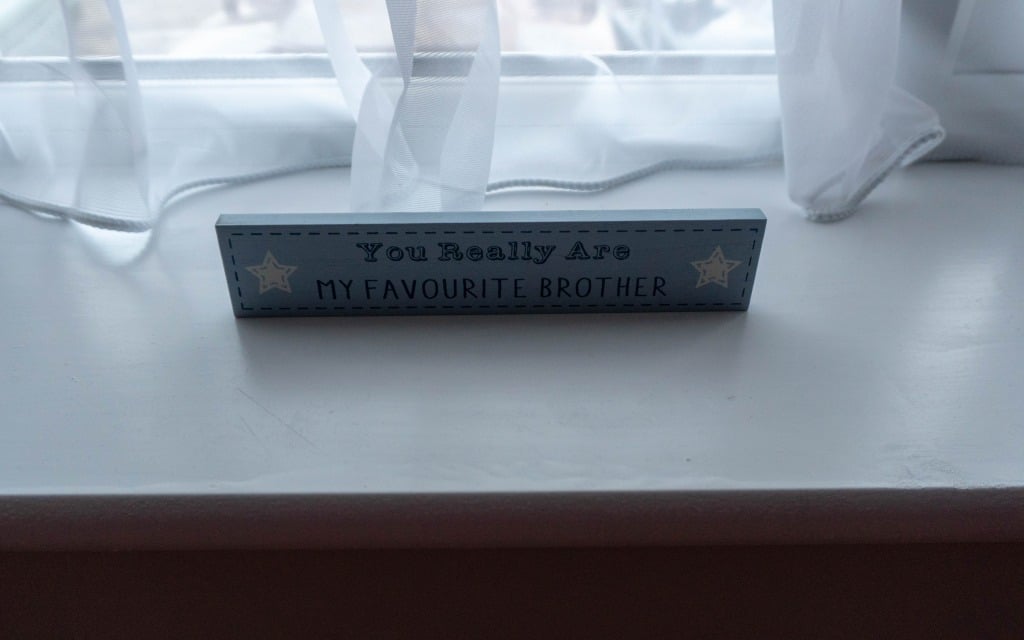 A light blue wooden sign on a windowsill reads, You Really Are MY FAVOURITE BROTHER with two white stars on each side—perfect for a teenage boy's bedroom or boys' navy bedroom. Sheer white curtains and part of the window are visible behind.