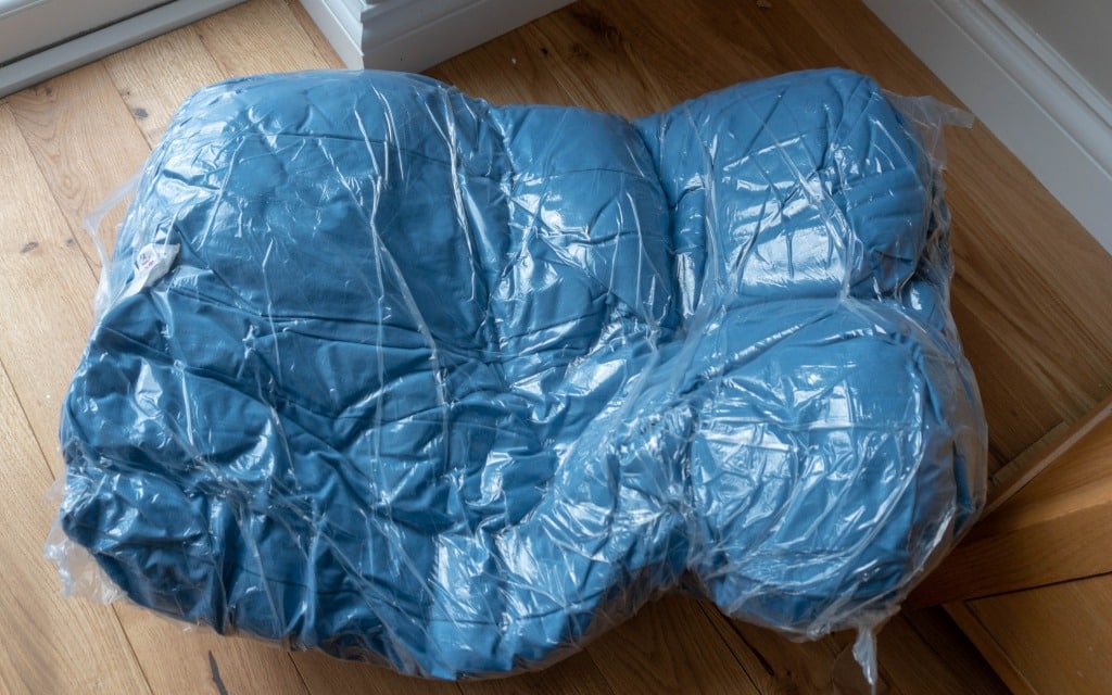 A blue Lupo's Nest Dog Bed is sealed inside a clear plastic bag, resting on a wooden floor near the corner of a room and a piece of furniture.