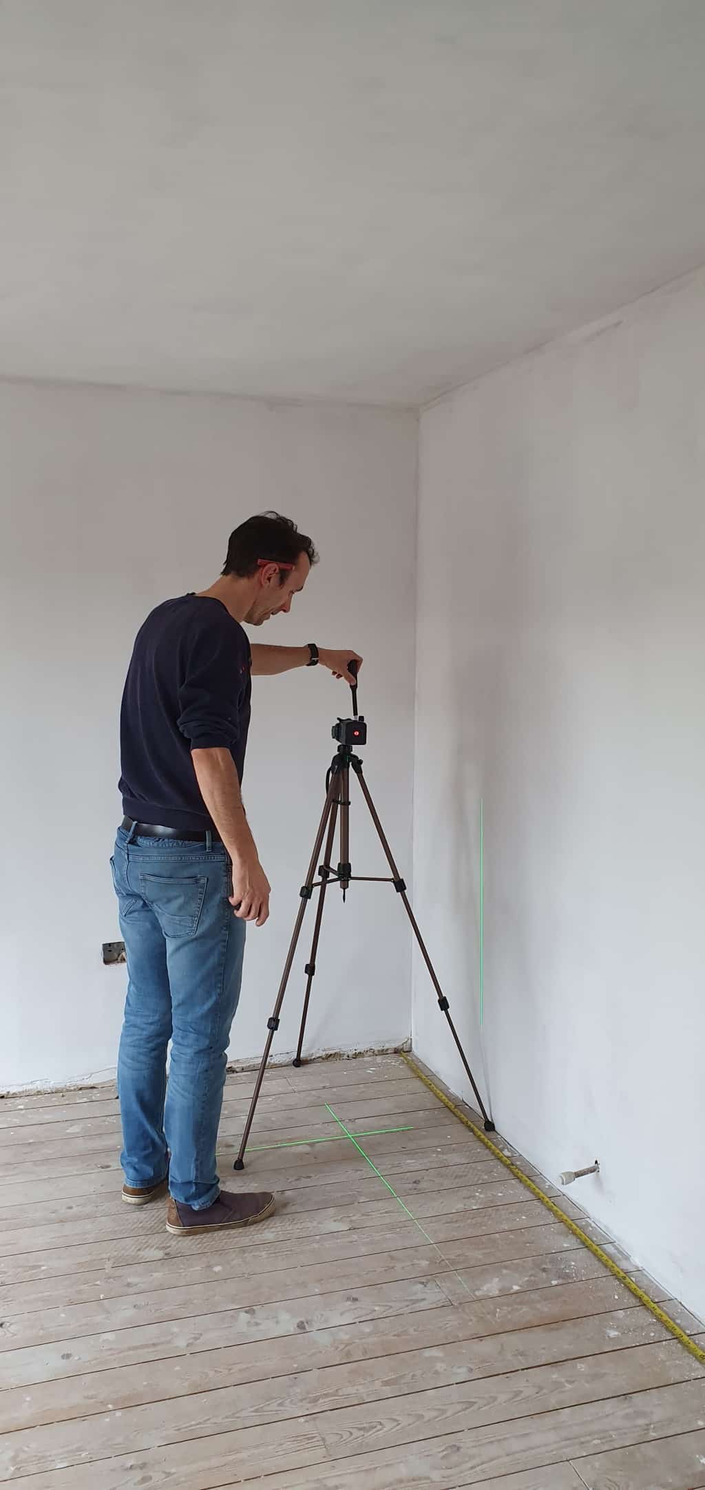 A man stands on a wooden floor in an unfinished room, adjusting a tripod with a laser level projecting green lines onto the white walls and floor during a family home renovation.