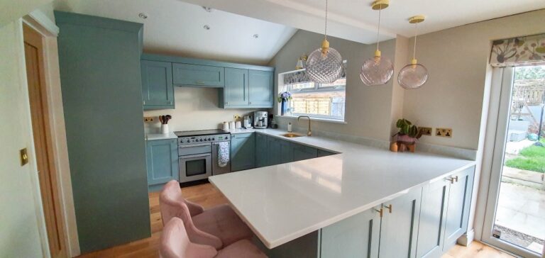 Modern blue and gold kitchen with light blue cupboards, a large white island worktop, two pink bar stools, and three pendant lights. Plants and large windows brighten this stunning Dining Room Reveal.