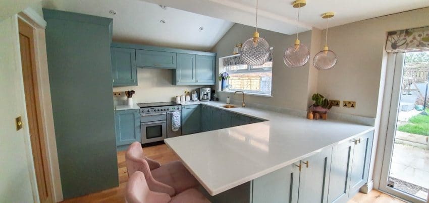 Modern blue and gold kitchen with light blue cupboards, a large white island worktop, two pink bar stools, and three pendant lights. Plants and large windows brighten this stunning Dining Room Reveal.