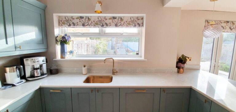 Bright, modern Blue and Gold Kitchen with light blue cupboards, white marble worktops, gold fittings, a coffee machine, and floral-patterned blinds above the sink that fill the space with natural light. Dining Room Reveal coming soon!.