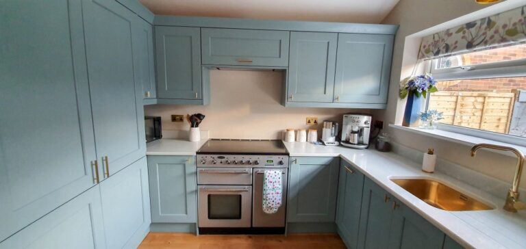 A modern Blue and Gold Kitchen with light blue cupboards, a stainless steel oven, and a gold sink. Sunlight streams through a window above the sink, decorated with flowers—an inspiring Kitchen Reveal full of charm.