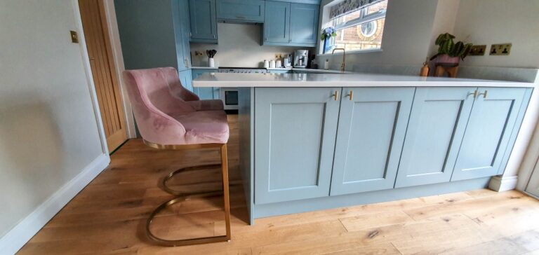 A modern blue and gold kitchen with light blue cupboards, a white worktop, and wooden flooring. Two pink velvet bar stools with gold legs sit at the island. A plant and kitchen appliances complete this stylish kitchen reveal.