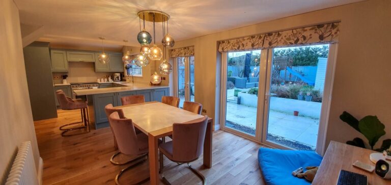 A modern dining area with a wooden table, pink chairs, and a cluster of hanging lights sits beside a kitchen with pastel cupboards. Large glass doors open to the patio—ideal for those seeking inspiration when choosing wall paint colours for a fresh look.