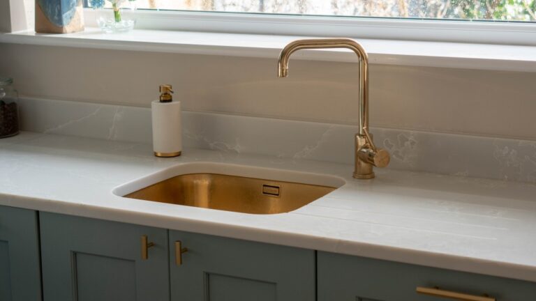 A modern Blue and Gold Kitchen worktop with a gold sink, matching gold tap, white soap dispenser, and blue cupboards sits beneath a large window filled with natural light.