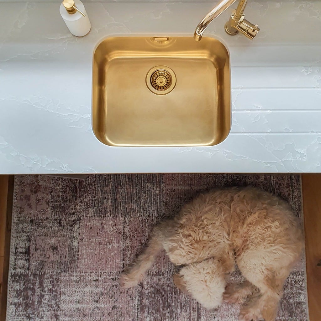 A fluffy, light-coloured dog is curled up on a patterned rug atop solid wood kitchen flooring beneath a modern white worktop with a gold-coloured sink and a soap dispenser. The image is taken from above.